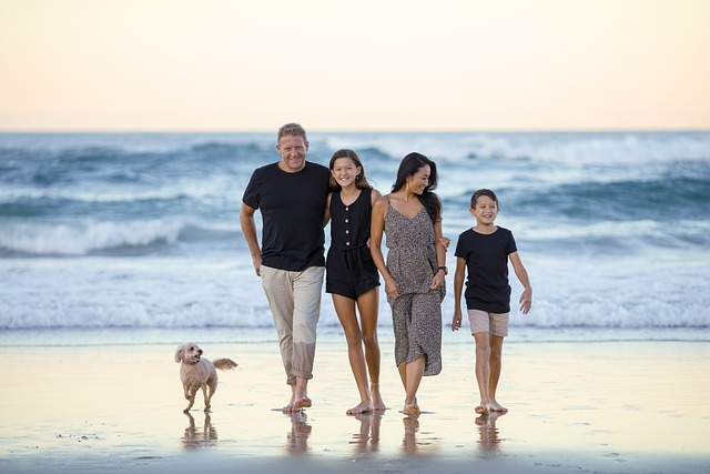 A happy family of four, consisting of a man, woman, teenage girl, and young boy, walks along the beach at sunset. The parents are holding hands with their children, all smiling and enjoying the moment. A small dog trots alongside them on the wet sand near the gentle waves of the ocean, creating a serene and joyful atmosphere.