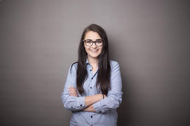 A young woman with long dark hair and glasses stands confidently with her arms crossed, wearing a light blue button-down shirt. She is smiling and appears relaxed, standing against a plain gray background, conveying a sense of professionalism and self-assurance.