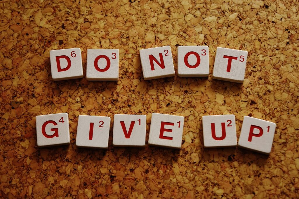 Letter tiles on a corkboard spelling out the motivational message "DO NOT GIVE UP." The tiles are arranged in two lines, emphasizing persistence and encouragement.