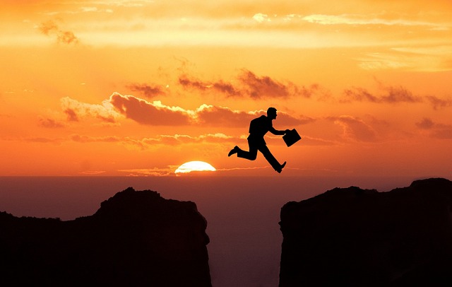 "Silhouette of a person with a briefcase leaping across a gap between two cliffs at sunset, symbolizing the determination and courage needed to overcome obstacles."